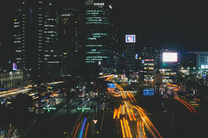 A busy cityscape with streaming light trails of cars whizzing by tall buildings at night