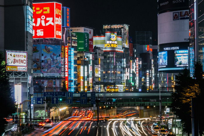 A busy Japanese cityscape with streaming light trails of cars whizzing by tall buildings