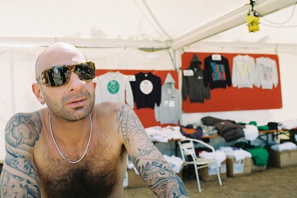 Photo of a topless man with arms covered in tattoos in sunglasses inside a merch tent