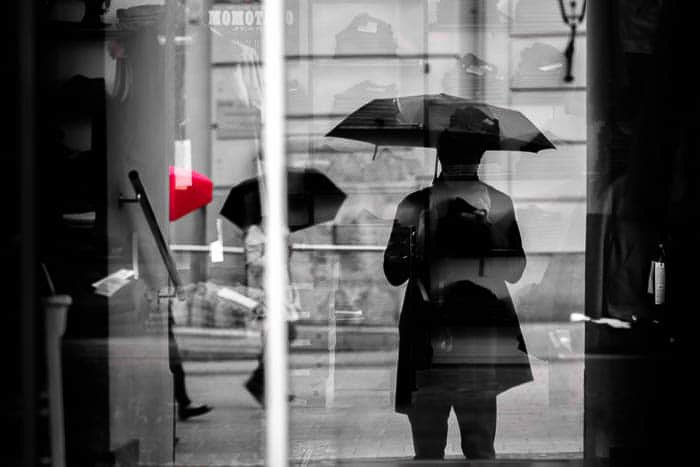 A black and White street photo of people walking in the rain, one umbrella is spot colored red 