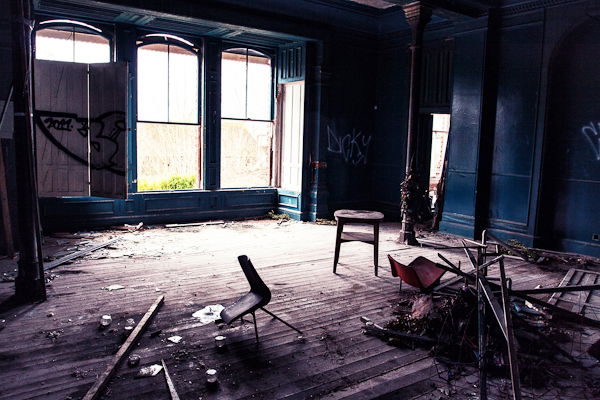 Room in an abandoned building with big windows, dark painted walls and broken wood and chairs