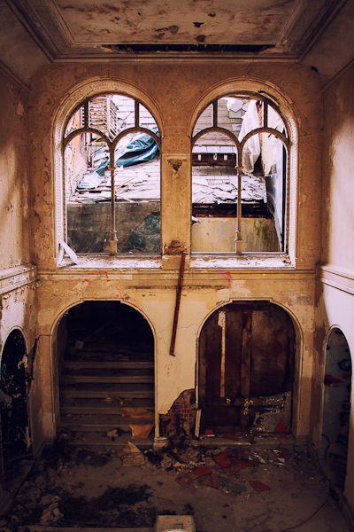 Abandoned building from the inside, two windows looking out to a ruined rooftop and a dark staircase leading up.