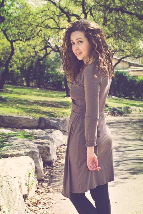 cropped photo of curly haired brunette in a brown dress and boots looking back while walking down a dirt path in a park