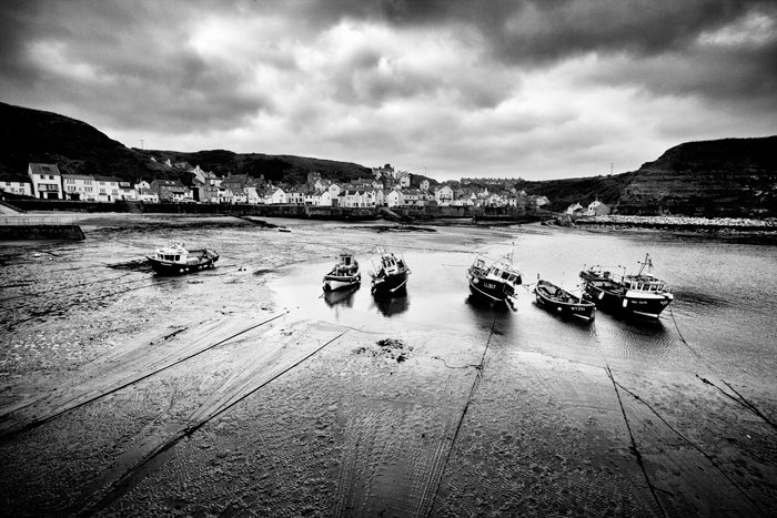A Black and white landscape photo of a line of boats sitting in harbour at low tide