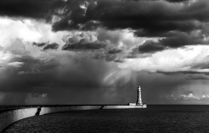 A stunning black and white shot of sun-kissed lighthouse on promontory with dark clouds overhead