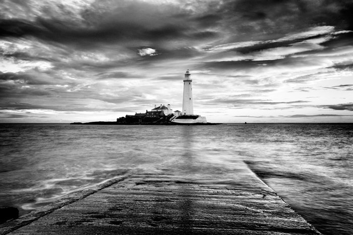 Black and white coastal view across the water looking towards a lighthouse, shot with a long exposure