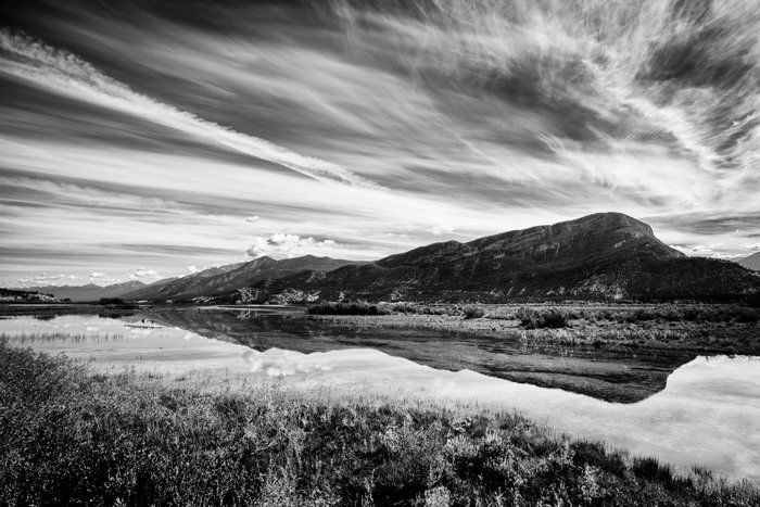 A black and white photo of a cloud streaked sky reflected in water with hill in background