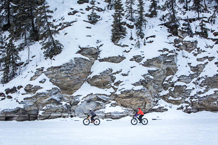 View from afar of two cyclists riding in the snow. The mountain side is in the background.