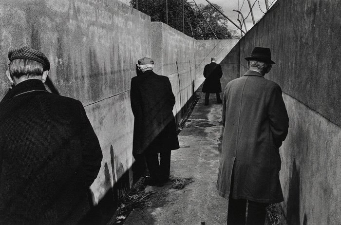 Black and white street photography of four men stand in a bleak corridor urinating 