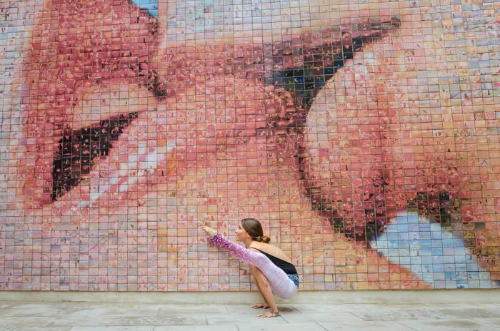 Artistic yoga photography of a model in firefly pose in front of a mosaic wall showing two people kissing