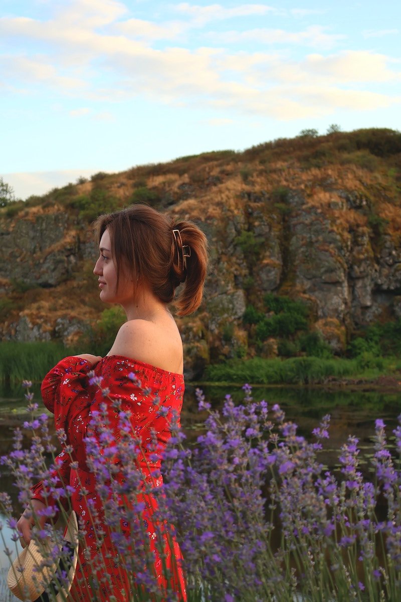 A woman standing by flowers in a side profile with trees in the background
