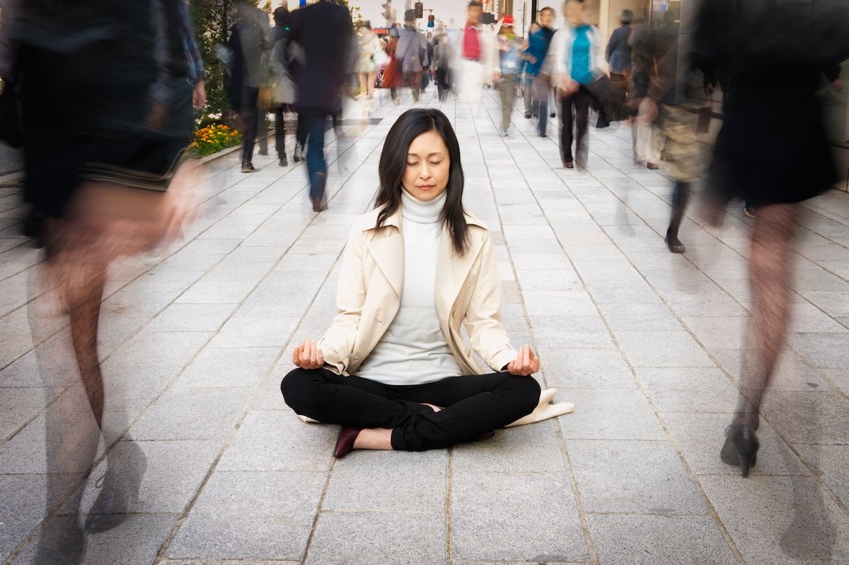 Yogini sitting on a sidewalk in a sitting pose for a long exposure with people walking by as an example of creative yoga photography