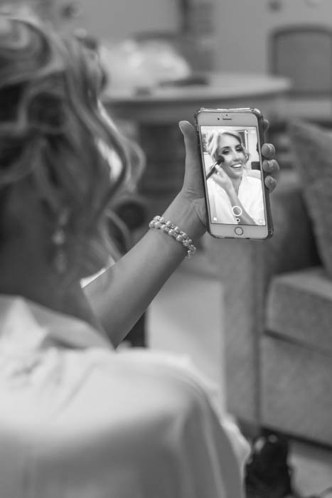 A black and white wedding photo of bride taking a selfie while doing makeup 