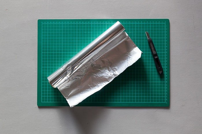 An overhead shot of a cutting mat, tin foil roll and Stanley knife - materials for how to make a diy lightbox for photography