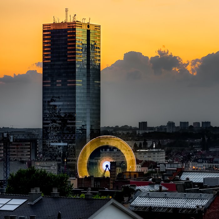 bright yellow sky and clouds behind tall buildings, rooftops and the yellow glow from the movement of a Ferris wheel.The result of a long exposure taken with a 10-stops ND filter.