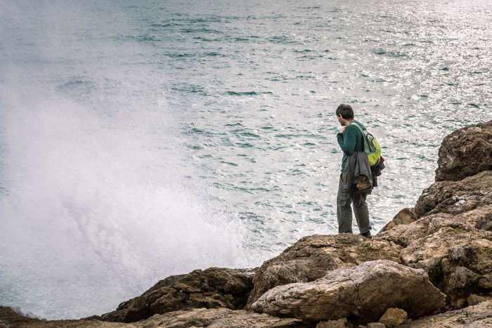 A lone figure standing on rocks by the beach and staring into the water