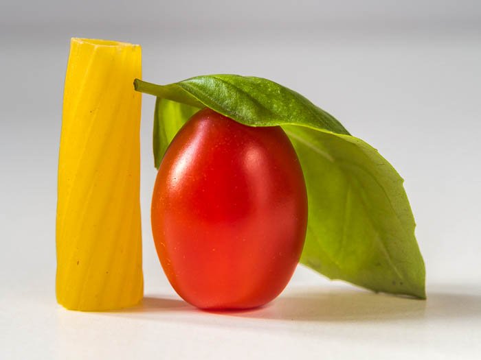 foto de comida minimalista cerca de una pieza de pasta rigatoni, un tomate ciruela y una hoja de albahaca
