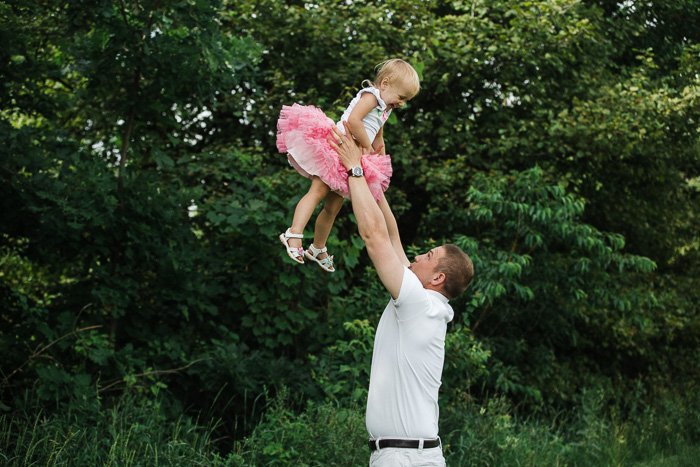 playful family portrait of a father holding a little girl in pink tutu above his head in a leafy outdoor setting