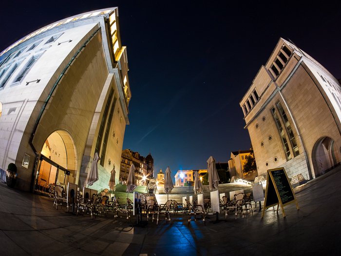 powerful distorted image of two tall buildings framing a plaza area of tables and chairs at night, urban photography taken with fisheye lens