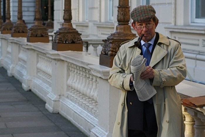 London gentleman in peaked cap and glasses, smoking a pipe and reading the newspaper in an outdoor urban setting