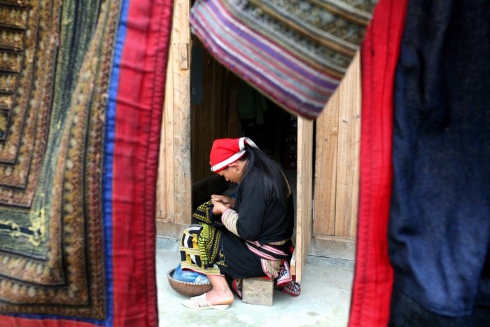 Travel photography showing a Vietnamese lady sitting down while working with textiles, framed by coloured materials hanging in the foreground