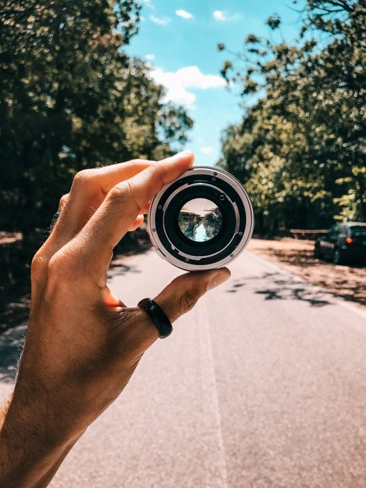 A hand holding up camera lens showing a city road with trees on the side