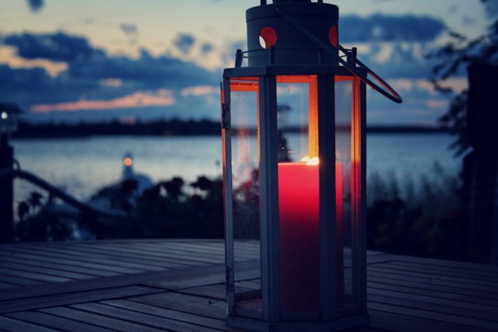 Red candle lit inside a lantern set on a wooden table next to a lake at dusk