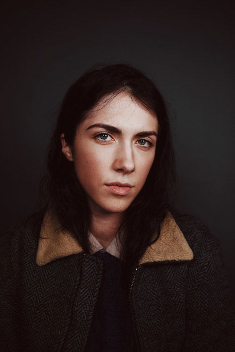 Serious female model looking into the camera, standing in front of a black background, wearing a sheep skin lined jacket