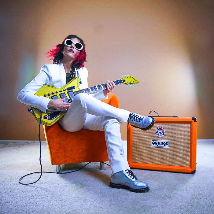 A woman sitting on an orange chair beside an orange amp holding an electric guitar