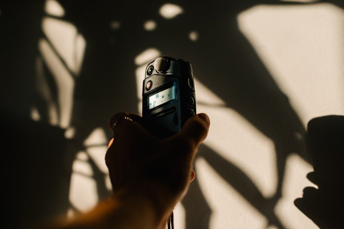 A hand holding a light meter up to an indoor wall with dappled light and shadows