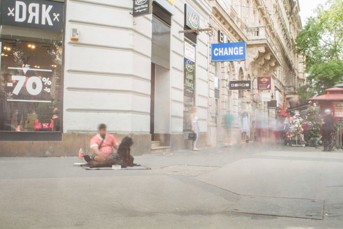 A long exposure street photograph in Budapest, a blurred homeless man sits on the ground with blurred figures in the background.