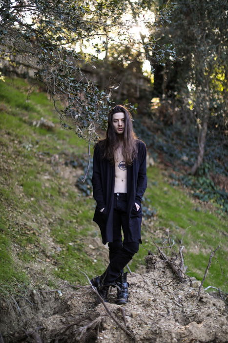 Portrait of a man with long hair, black clothes and tattooed chest, standing in a forest area, looking at the camera. Improve your photography skills today.