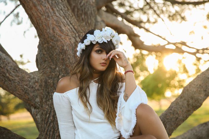 Bright and airy portrait of a girl in white dress with white flowers in her hair, sitting in front of a tree. Improve your photography skills today