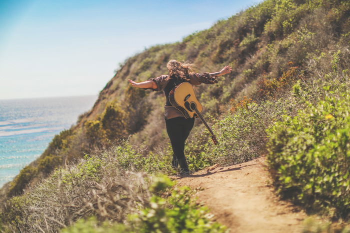 Outdoor photo of a person with acoustic guitar on their back, walking away from the camera on a coastal path, arms outstretched. Improve your photography skills today.