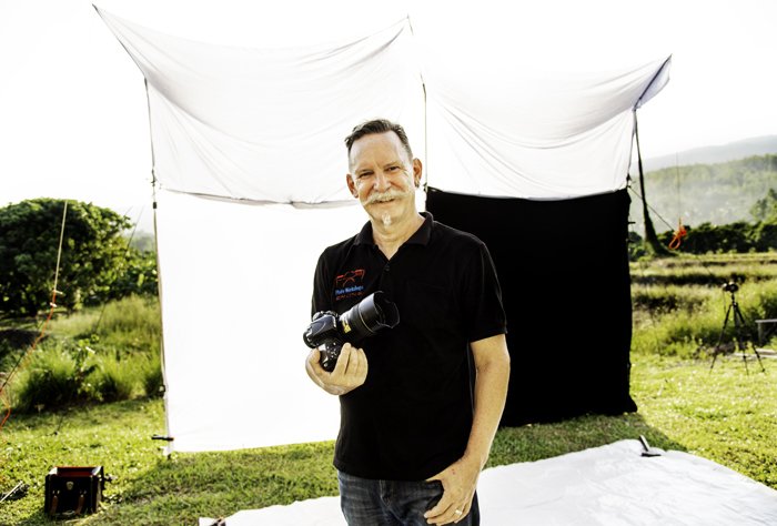 A photographer poses with his camera in front of his outdoor photography studio. 