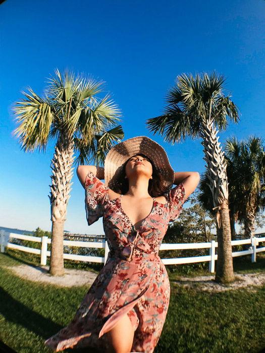 Girl in pink flowery dress and straw hat standing on the grass in front of a white fence and palm trees, holding her arms behind her head on a bright day - Smartphone fashion photography shoot