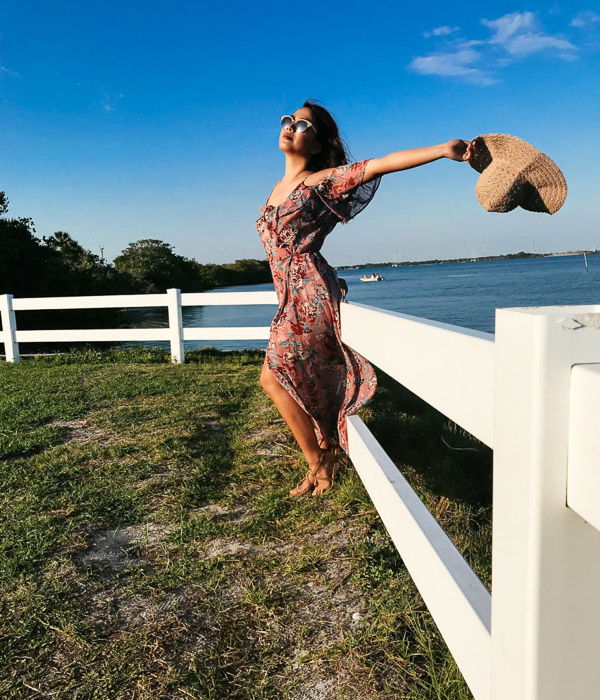  Girl in pink flowery dress and straw hat leaning against a white fence by the sea, holding her hat behind her head on a bright day - Smartphone fashion photography shoot