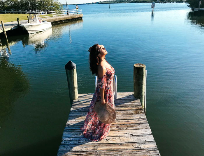 Girl in pink flowery dress and straw hat standing on a wooden dock by the sea, holding her hat behind by her side on a bright day - Smartphone fashion photography shoot