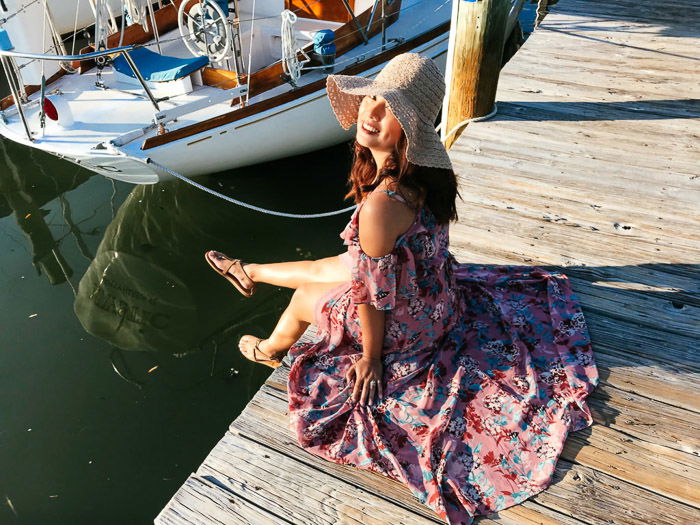 A girl in flowery dress and straw hat sits on a dock beside a boat, smiling at the camera. Location is important for Smartphone fashion photography