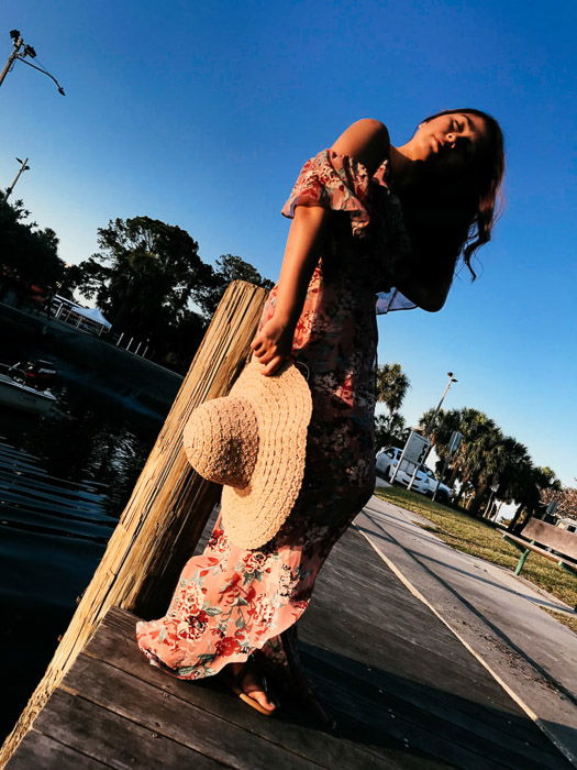 Girl in pink flowery dress on a wooden dock by the sea, holding her hat by her side on a bright day - Smartphone fashion photography shoot