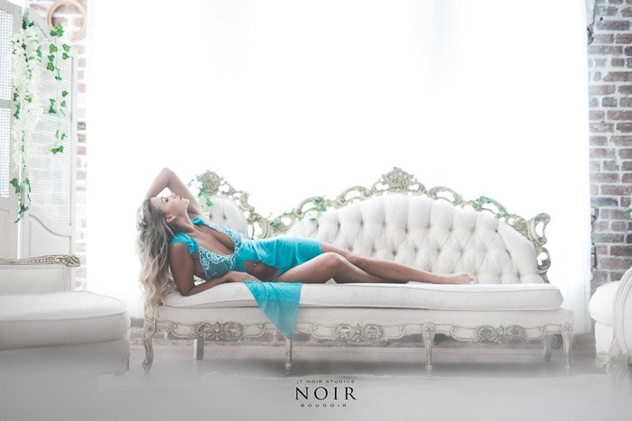 Light and airy boudoir shoot of a blonde girl in turquoise dress lying back on a white chaise lounge