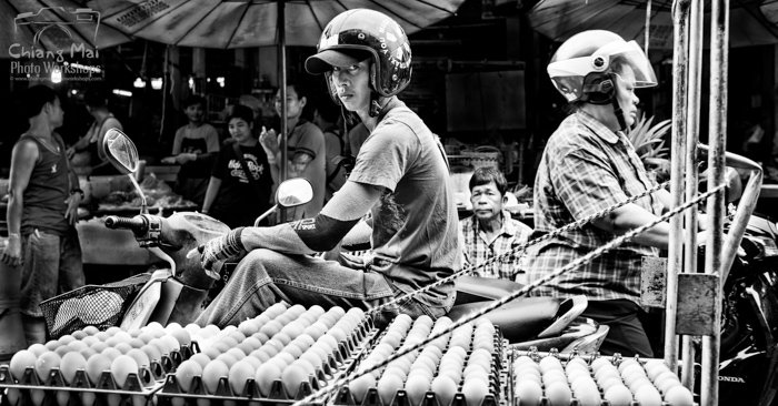 Black and white documentary photography of men on motorbikes riding through a busy market in Chiang Mai, Thailand