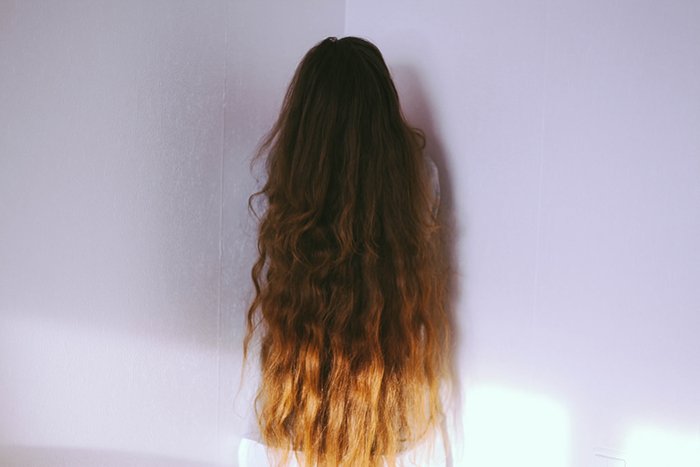 一个女孩用她长长的棕色头发遮住了她的脸，站在一个粉红色的房间里，摆姿势拍一张无脸的肖像。