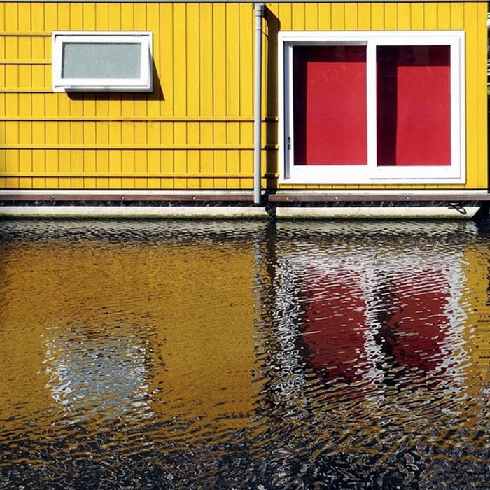 Photo of a yellow house with red windows reflected in the water below 