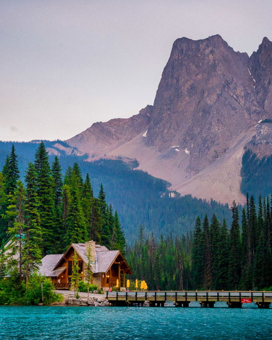 Photo of a wooden house by a bridge over blue water with a mountainous background. Landscape photography composition