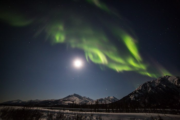 Photo of the moon lighting up the Brooks Range of northern Alaska in warm light with the aurora borealis above. 
