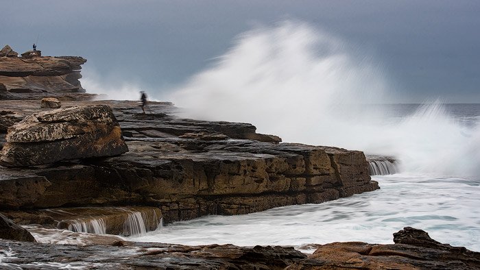 Seascape photo of a two people fishing on the rocks with large waves behind.