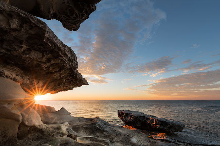 Blended sunburst seascape photo with rocks and cloudy blue sky.