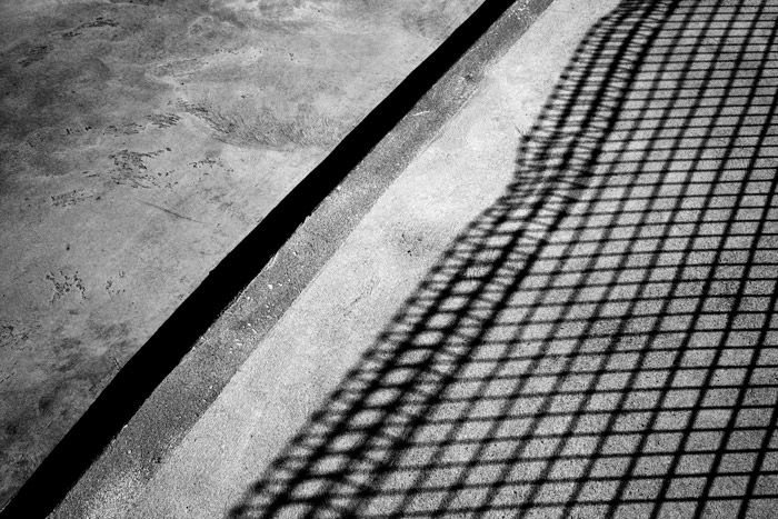 A shadowy black and white photograph of concrete with shadow of a fence. 