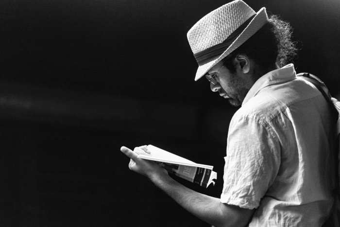 Black and white candid photograph of a man in a white hat and shirt reading a book